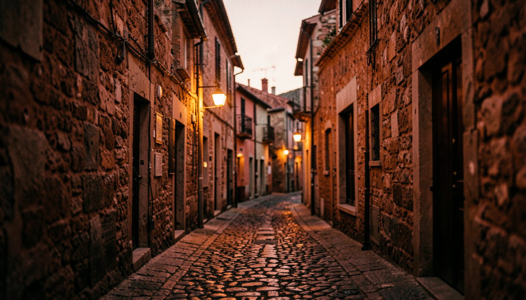 Narrow cobblestone street in an Umbrian hill town at dusk, warm terracotta and stone tones, soft lantern light